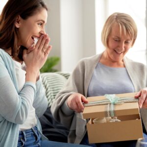 Daughter watching with delight as mother opens gift on a couch in a brightly sunlit room.
