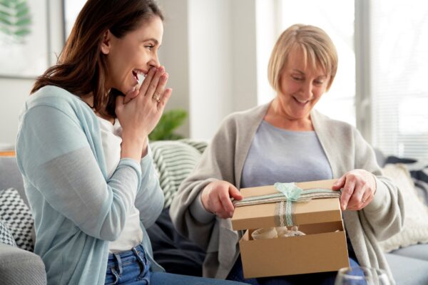 Daughter watching with delight as mother opens gift on a couch in a brightly sunlit room.
