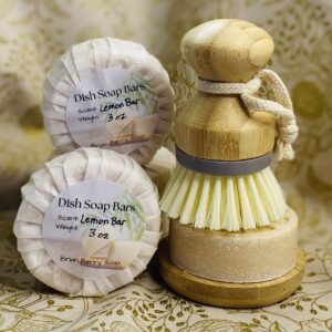 Dish soap bars wrapped and labeled and one bar unwrapped resting on a soap dish with a scrub brush on top.