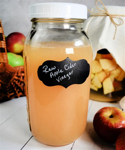 Apple cider vinegar in a mason jar surrounded by apples in a basket and on a whitewashed counter top with a fresh batch of vinegar brewing in the background.